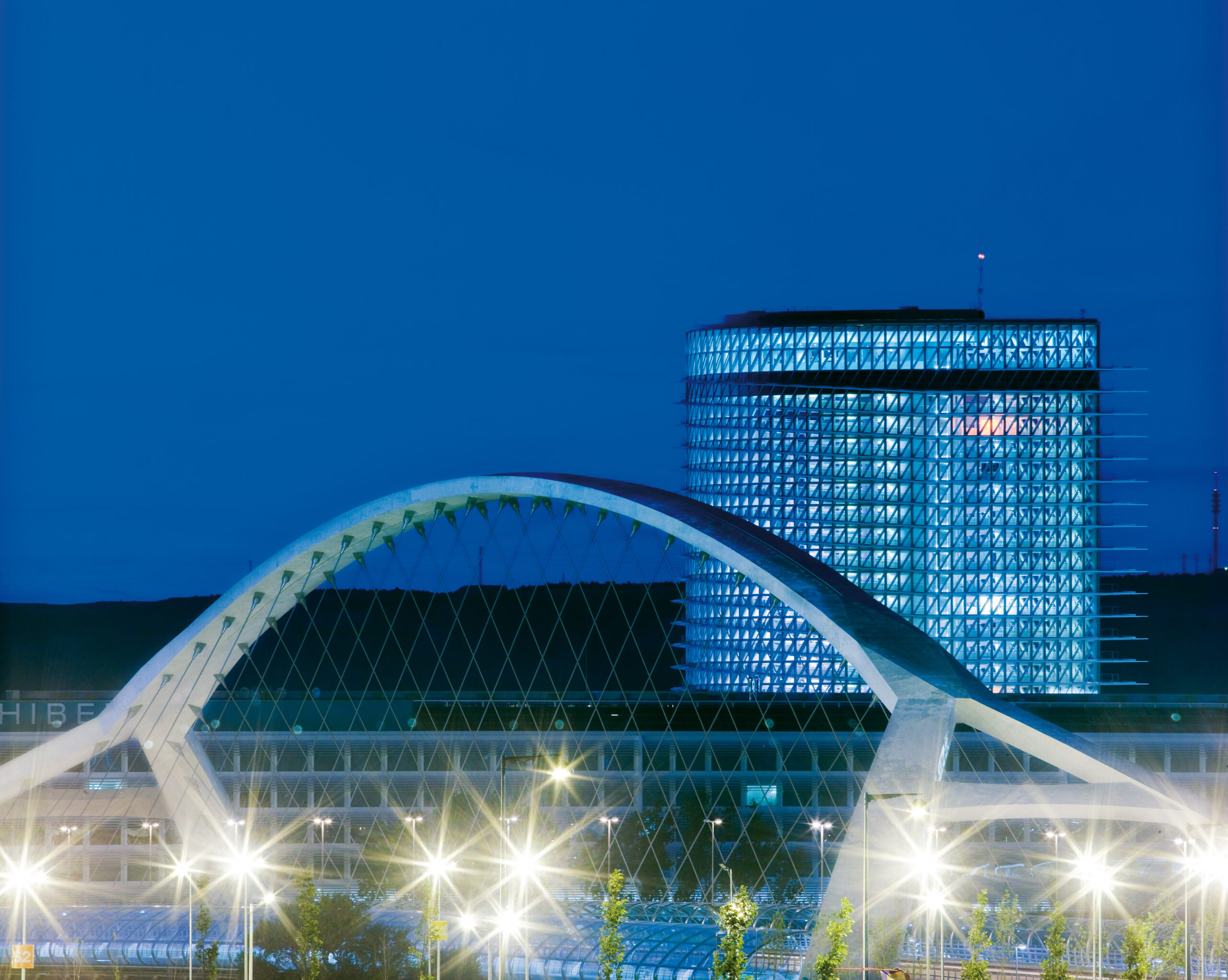 Vista nocturna detallada que muestra el arco superior de hormigón blanco del Puente del Tercer Milenio cruzando diagonalmente frente a la Torre del Agua iluminada en azul, con brillantes luces de farolas en la parte inferior creando destellos en forma de estrella.