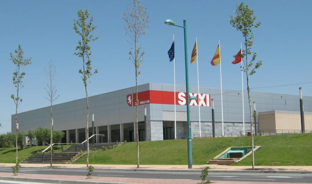 Fachada del Centro Deportivo Municipal Siglo XXI con banderas oficiales y zona ajardinada en primer plano.