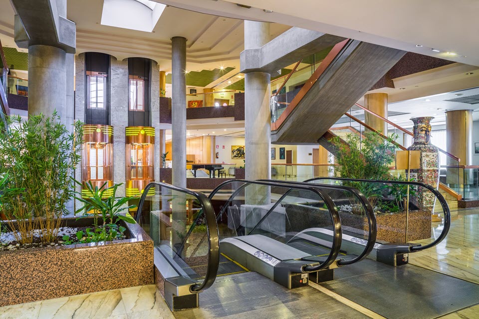 Vestíbulo del Hotel Exe Boston Zaragoza con escaleras mecánicas, columnas de hormigón y zonas ajardinadas interiores.