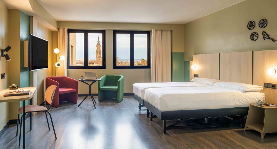 Habitación luminosa del Hotel Ibis Zaragoza Centro con cama doble, sillones de colores y vistas a la basílica del Pilar.